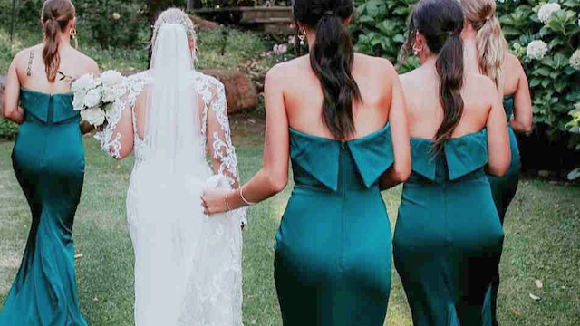 Noiva caminhando num jardim com quatro damas de honor vestidas com vestidos longos de cetim na cor verde esmeralda, com decote sem alças e detalhe de laço nas costas, criando um visual elegante e coordenado.