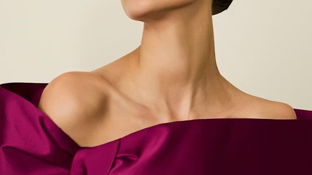 Close-up do decote de um vestido de festa em cetim estruturado na cor magenta (vinho/púrpura). O design destaca um corte sofisticado de ombro a ombro assimétrico, evidenciando o colo e a clavícula da modelo. O fundo é neutro e minimalista.