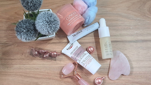 Produtos de skincare sobre uma mesa de madeira, incluindo o Clinique Moisture Surge, o gel de limpeza Eucerin Anti-Pigment, o sérum Endocare Age Barrier, o protetor solar Medik8 e ferramentas de massagem facial em quartzo rosa.