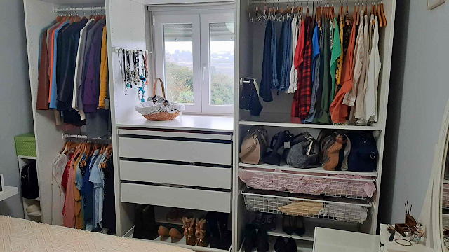 Fotografia de um closet organizado com roupa pendurada, gavetas brancas, sapatos arrumados em prateleiras e luz natural a entrar pela janela ao centro.