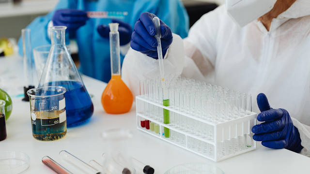 Imagem em close-up focada nas mãos enluvadas de azul de dois pesquisadores num ambiente de laboratório. No primeiro plano, um cientista com um fato de proteção branco e máscara usa uma pipeta para transferir uma substância líquida de cor verde para um tubo de ensaio dentro de uma prateleira de tubos cheia. A mão direita do cientista segura a pipeta, enquanto a mão esquerda, também com luva azul, segura a base da prateleira. No fundo desfocado, outro cientista com um fato de proteção azul manipula um objeto pequeno e não identificado. O laboratório está repleto de equipamentos de vidro, como balões e béqueres, cheios de líquidos coloridos.