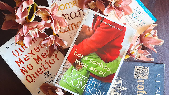 "Livros dispostos sobre uma mesa escura com flores cor-de-rosa, incluindo os títulos 'Quem Mexeu no Meu Queijo?', 'Comer, Orar, Amar', 'Bons Sonhos, Meu Amor' de Dorothy Koomson, 'A Guardadora de Histórias' de Jodi Picoult e 'Profecia' de S.J. Parris.