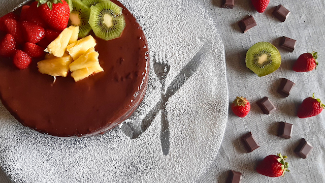 Bolo de chocolate decorador com morangos, Kiwi, e ananás sobre um parto decorado com açúcar em pó. Ao lado, quadrados de chocolate, morangos e kiwis.