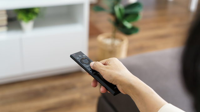 Mão a segurar um comando de televisão enquanto a pessoa está sentada no sofá, com o ambiente da sala desfocado ao fundo.