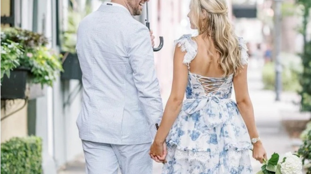 A couple walks hand-in-hand outdoors, showcasing elegant garden wedding fashion. The woman is wearing a blue and white floral dress with lace trim and a corset-style back. The man is wearing a light blue seersucker summer suit. The aesthetic is romantic, classic, and perfect for an outdoor ceremony.