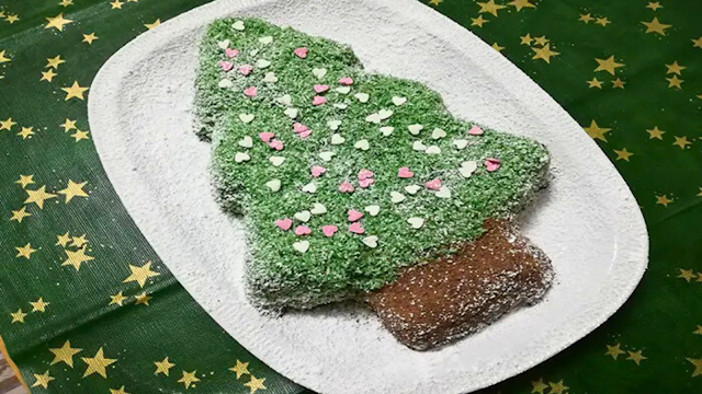 Bolo de Natal em forma de pinheiro decorado com coco colorido Bolo de Natal em forma de árvore de Natal, coberto com coco ralado verde e decorado com pequenos corações em açúcar, servido num prato branco sobre toalha verde com estrelas douradas