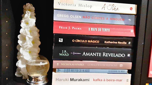 Pilha de Leituras Favoritas em Prateleira Decorada Fotografia em close de uma pilha vertical de oito livros deitados em uma prateleira escura. À esquerda, há um objeto decorativo branco em formato de árvore de flores e um pequeno globo de neve. As lombadas dos livros exibem títulos como "Kafka à Beira-Mar" de Haruki Murakami, "Amante Revelado" de J.R. Ward e "A Ilha" de Victoria Hislop.
