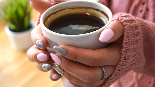 Plano aproximado de uma pessoa a segurar uma caneca branca com café preto entre as mãos. A pessoa veste uma camisola de malha cor-de-rosa e exibe uma manicure cuidada com unhas em tons de rosa claro, cinzento e prateado brilhante. Ao fundo, vê-se o detalhe desfocado de uma planta verde, criando um ambiente acolhedor e caseiro.