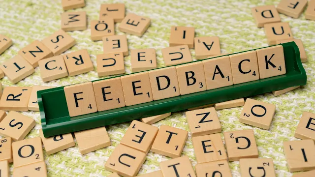 Peças de um jogo de palavras cruzadas (estilo Scrabble) organizadas em um suporte verde para formar a palavra "FEEDBACK". O suporte está posicionado sobre um fundo repleto de outras peças de letras espalhadas de forma aleatória em tons de madeira clara.