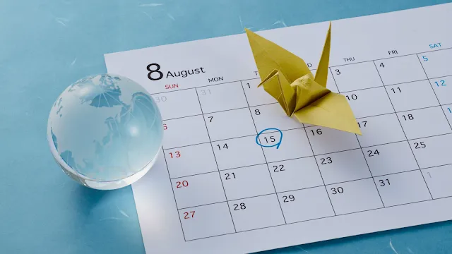 Calendário de parede aberto no mês de agosto sobre um fundo azul claro. Do lado esquerdo, há um pequeno globo terrestre de vidro transparente. Em cima do calendário, cobrindo os dias 2, 3, 9 e 10, destaca-se um tsuru (pássaro de origami) feito de papel dourado. O dia 15 está circulado com uma caneta azul.