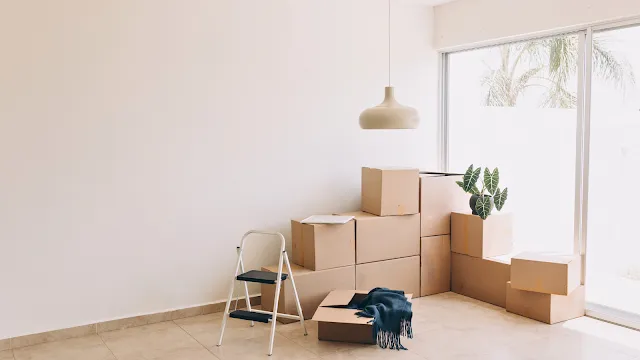O recomeço de uma nova mudança. Sala de estar vazia e iluminada durante um processo de mudança de casa. No centro, há várias caixas de papelão empilhadas perto de uma grande janela de vidro que dá para o exterior. Em primeiro plano, vê-se um pequeno escadote branco e uma caixa aberta com uma manta azul escura. Uma planta em vaso decora o topo de uma das caixas sob um candeeiro suspenso.