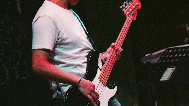 Fotografia em plano médio e de perfil de uma pessoa a tocar um baixo elétrico num palco escuro. A pessoa veste uma t-shirt branca simples e o baixo tem um corpo preto com um escudo branco e uma cabeça vermelha. A mão esquerda pressiona as cordas no braço do instrumento, enquanto a mão direita está posicionada sobre as cordas perto dos captadores. O ambiente tem uma iluminação contrastante com tons avermelhados, e ao fundo, do lado direito, é possível ver parte de um suporte com uma pauta ou tablet.
