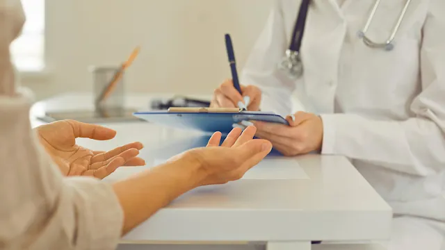 Uma fotografia de close-up tirada ao nível da mesa mostra as mãos de um paciente estendidas em um gesto expressivo em direção a uma médica do SNS. A médica, vestida com um jaleco branco e com um estetoscópio em torno do pescoço, está sentada à direita e toma notas ativamente em uma prancheta azul com uma caneta. O plano de fundo é embaçado, sugerindo um ambiente clínico luminoso e tranquilo. Esta imagem evoca a interação humana e o cuidado compassivo no coração do Serviço Nacional de Saúde português.  Esta opção conecta a imagem diretamente ao contexto do SNS e do seu atendimento "Clássico Atual e Normal".