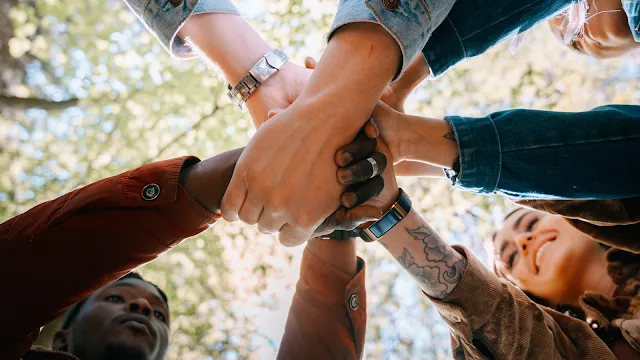 Uma perspectiva de baixo para cima mostra um grupo diverso de jovens unindo as mãos ao centro, formando um círculo de apoio. É possível ver detalhes como tatuagens, relógios e diferentes tons de pele sob a luz do sol que filtra pelas copas das árvores ao fundo. Rostos sorridentes aparecem parcialmente nas bordas, transmitindo um sentimento de coletividade e amizade em um ambiente externo.