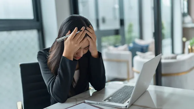 Mulher jovem, de cabelo escuro e vestindo um blazer preto, sentada numa mesa de escritório em frente a um computador portátil aberto. Ela está com a cabeça baixa, os olhos fechados e segura o rosto com as duas mãos, demonstrando uma expressão de forte dor, stress ou enxaqueca. Na mão direita, segura uma caneta. O fundo do ambiente de trabalho está ligeiramente desfocado.