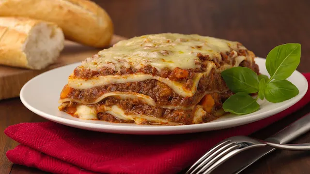 Uma fatia quadrada de lasanha caseira servida em um prato branco, revelando camadas bem definidas de massa, molho à bolonhesa com carne moída e molho branco. O topo está coberto por uma camada generosa de queijo muçarela derretido e levemente dourado. Ao lado, há um ramo de manjericão fresco e, em primeiro plano, talheres de metal sobre um guardanapo de tecido vermelho. Ao fundo, pedaços de pão francês repousam sobre uma tábua de madeira desfocada.