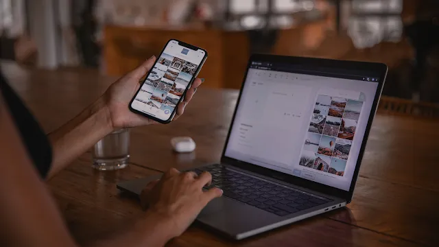 Criador de conteúdo a planear publicações de um blog no portátil e telemóvel. Uma pessoa segura um smartphone na mão esquerda, mostrando uma grelha de fotografias estéticas, enquanto a mão direita digita no teclado de um portátil MacBook Pro aberto numa mesa de madeira escura. O ecrã do portátil exibe uma plataforma de edição e organização de conteúdos visuais. O ambiente ao fundo está desfocado.