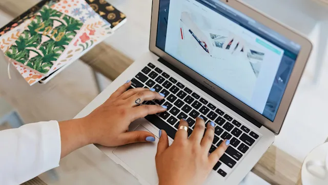Vista superior das mãos de uma pessoa com unhas pintadas de azul e uma tatuagem de coração no dedo, a digitar no teclado de um portátil MacBook Air. O ecrã mostra uma imagem de alguém a desenhar ou a escrever. Ao lado, sobre a mesa de vidro, destacam-se cadernos com capas coloridas e padrões florais.