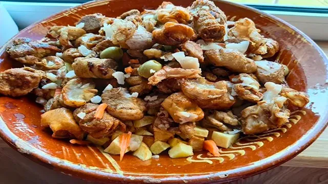 Prato Tradicional de Rojões à Moda do Porto em Travessa de Barro Uma travessa redonda de barro vidrado, decorada com padrões tradicionais, cheia até ao topo com rojões à moda do Porto. O prato inclui cubos de carne de porco frita bem dourados, batatas em cubos no fundo, tripas enfarinhadas, pedaços de carne e pickles cortados, decorado com azeitonas verdes inteiras. A imagem é tirada de um ângulo ligeiramente superior, destacando a textura rica e o aspeto reconfortante da comida caseira.