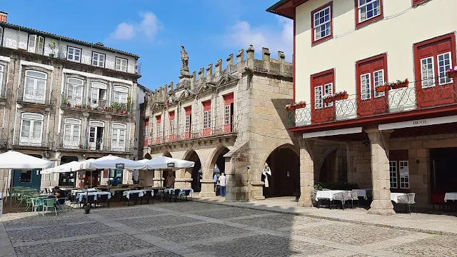 Vista da Praça da Oliveira, em Guimarães, Portugal, num dia ensolarado. Ao centro, destaca-se o Antigo Paço do Concelho, um edifício medieval de pedra com arcos góticos no rés-do-chão e ameias no topo. À esquerda e à direita, encontram-se edifícios históricos tradicionais com varandas de ferro forjado e portadas coloridas. No piso térreo, esplanadas de restaurantes com mesas e guarda-sóis brancos ocupam o chão de calçada portuguesa, com algumas pessoas a caminhar e sentadas. O céu está azul com nuvens ligeiras.