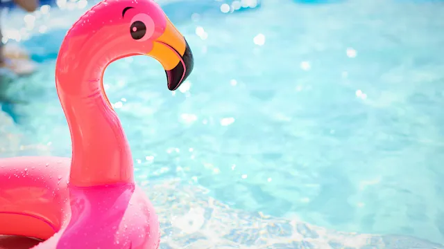 Vibrações de Verão: Mergulhos e Diversão na Piscina Primeiro plano de uma boia insuflável cor-de-rosa em forma de flamingo, com gotas de água na superfície, a flutuar numa piscina de água azul cristalina e cintilante sob a luz do sol.