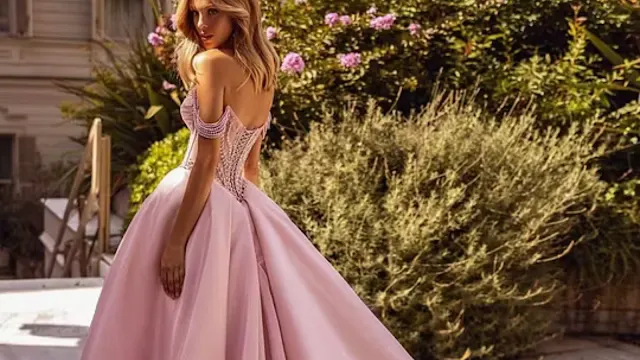 Vestido de Baile Rosa com Aplicações de Pérolas Mulher loira a usar um elegante vestido de baile rosa com corpete decorado com pérolas e alças descaídas, posando ao ar livre num jardim ensolarado.