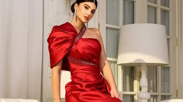 Uma mulher elegante com cabelo escuro preso e brincos longos, posando num interior luminoso. Ela usa um vestido de baile vermelho vibrante, de um ombro só, com detalhes drapeados exagerados no ombro e um cinto de pedraria. Ao fundo, vêem-se portas de vidro e um candeeiro de pé branco.