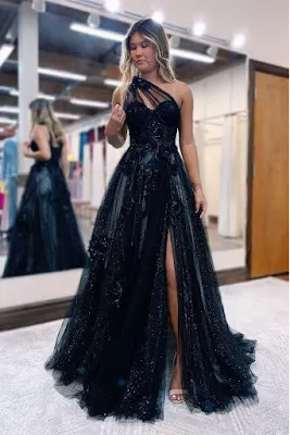 Blonde young woman posing in a fitting room wearing a long black prom dress with a one-shoulder design, shimmering tulle fabric, and a deep side slit.