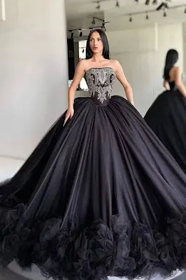 Uma mulher de cabelo preto comprido está em um provador e usa um vestido de baile preto extravagante com um corpete sem alças, detalhadamente bordado em prata e pedrarias, e uma saia de tule em camadas muito volumosa que termina com uma barra de babados. O reflexo dela é visível em um espelho ao fundo.