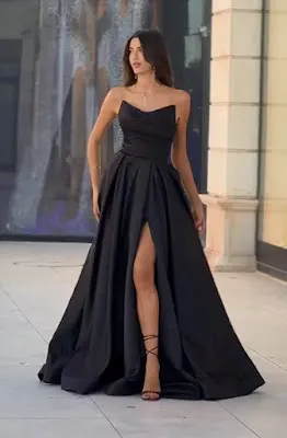 A tall woman with long brown hair poses outdoors wearing an elegant strapless black evening dress. The dress features a draped sweetheart neckline bodice and a full satin skirt with a high, bold leg slit. She completes the look with black strappy sandals and a delicate necklace. The background shows a store facade with a reflection.