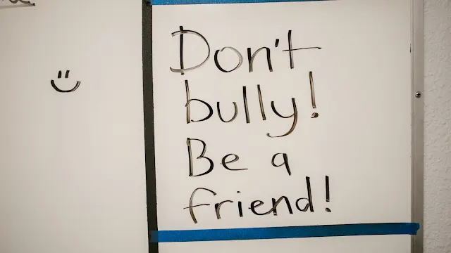 Fotografia aproximada de um quadro branco onde está escrita à mão, com marcador preto, a frase "Don't bully! Be a friend!". À esquerda da frase, visível no painel ao lado, encontra-se um pequeno emoji sorridente desenhado.