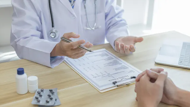 Consulta médica sobre reumatologia Médico com bata branca e estetoscópio ao pescoço, sentado numa secretária de madeira, a conversar com um paciente durante uma consulta médica, gesticulando com uma caneta sobre documentos de saúde. Na mesa encontram-se também frascos de medicamentos e uma embalagem de comprimidos.