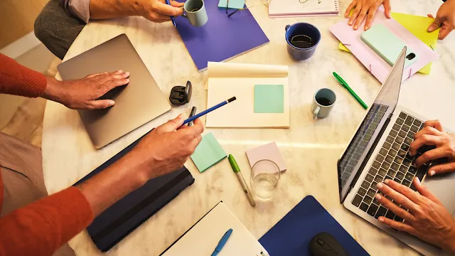 Reunião de trabalho colaborativa Vista superior de uma mesa redonda onde várias pessoas colaboram numa reunião de trabalho. A mesa está repleta de portáteis, cadernos abertos, blocos de notas autocolantes coloridos, canetas, chávenas de café e pastas de documentos, com as mãos dos participantes visíveis a interagir com os materiais.