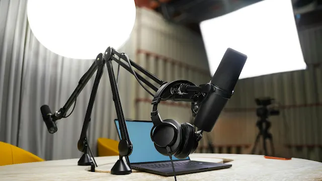 Dois braços articulados pretos com microfones profissionais e auscultadores pendurados estão instalados numa mesa de madeira clara, tendo ao fundo um computador portátil, luzes de estúdio difusas e uma câmara num tripé.
