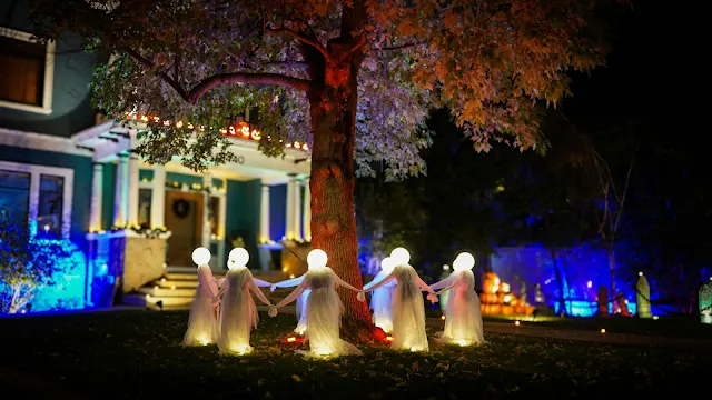Decoração de Halloween com figuras iluminadas em redor de uma árvore à noite Fotografia noturna de uma decoração de Halloween num jardim. Várias figuras humanoides com túnicas brancas compridas e cabeças em forma de esfera luminosa estão de mãos dadas a dar a volta a uma árvore de tronco largo. Ao fundo, vê-se uma casa iluminada com luzes azuladas e abóboras decorativas.