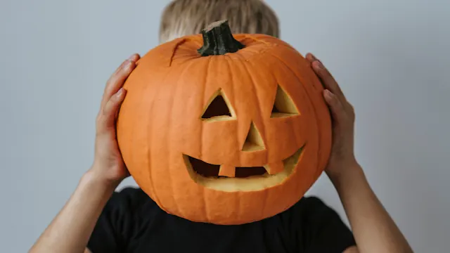 Criança com uma camisa preta segurando uma grande abóbora de Halloween esculpida com olhos triangulares e um sorriso largo à frente do rosto, cobrindo a cabeça contra um fundo cinza claro.