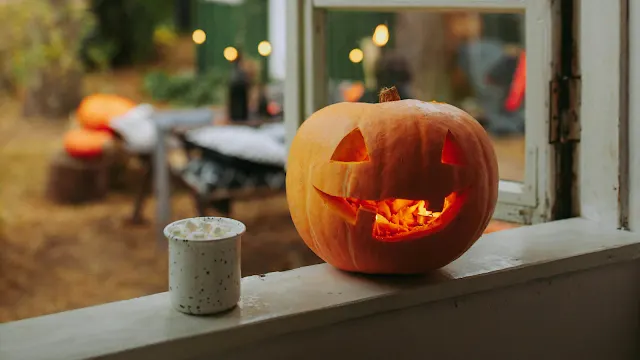 Abóbora de Halloween esculpida com rosto iluminado por dentro, posicionada no parapeito de uma janela, ao lado de uma caneca branca com marshmallows, tendo um cenário de outono desfocado ao fundo.